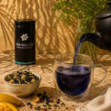 Tea being poured into a glass mug with tea leaves and a container labeled 'The Irie Cup' on a wooden surface.