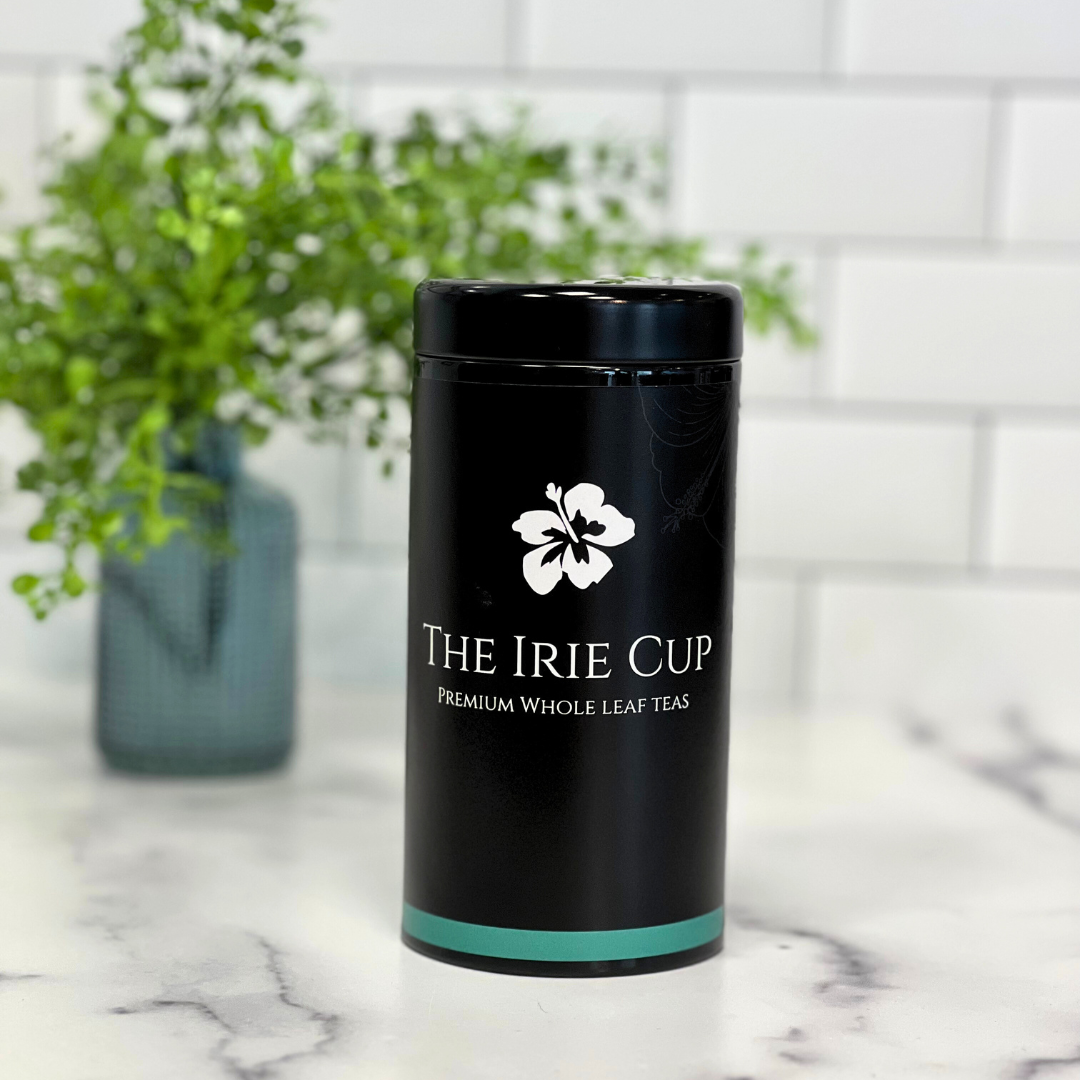A black cylindrical tea tin with a white floral logo and the text 'THE IRIE CUP Premium Whole Leaf Teas' on it, placed on a kitchen countertop with a white subway tile background and a green potted plant in the background.