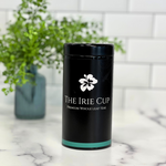 A black cylindrical tea tin with a white floral logo and the text 'THE IRIE CUP Premium Whole Leaf Teas' on it, placed on a kitchen countertop with a white subway tile background and a green potted plant in the background.