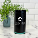 A black cylindrical tea tin with a white floral logo and the text 'THE IRIE CUP Premium Whole Leaf Teas' on it, placed on a kitchen countertop with a white subway tile background and a green potted plant in the background.