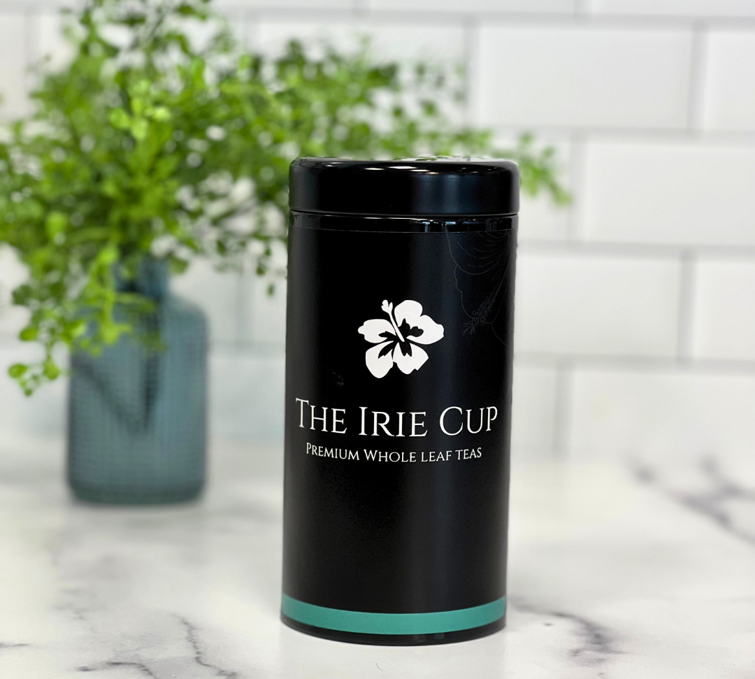 A black cylindrical tea tin with a white floral logo and the text 'THE IRIE CUP Premium Whole Leaf Teas' on it, placed on a kitchen countertop with a white subway tile background and a green potted plant in the background.