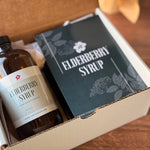 An elderberry syrup kit packaging with a bottle labeled 'Elderberry Syrup' and various herbs, placed on a wooden surface.