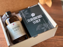 An elderberry syrup kit packaging with a bottle labeled 'Elderberry Syrup' and various herbs, placed on a wooden surface.