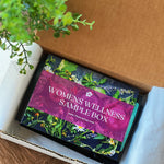 An open box labeled 'Women's Wellness Sample Box' with various herbal tea bags visible inside. The box is placed on a wooden surface with a green plant in the background.