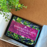 An open box labeled 'Women's Wellness Sample Box' with various herbal tea bags visible inside. The box is placed on a wooden surface with a green plant in the background.
